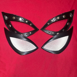 Op de afbeelding: Zwarte en witte oogstukken van een superheldenmasker. De oogstukken hebben een zwart frame met zilveren klinknagels en witte mesh ooglenzen. De maskeronderdelen zijn geplaatst tegen een rode achtergrond.