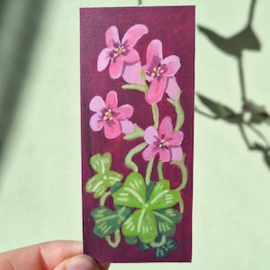 May include: A small rectangular painting of pink flowers with yellow centres and green leaves on a dark red backdrop. The artwork is held by a hand, with a light green background.
