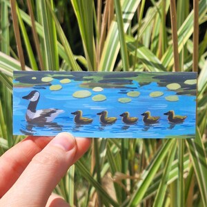 May include: A rectangular bookmark featuring a painted illustration of a Canada goose swimming in blue water followed by five small yellow ducklings. The scene includes scattered green lily pads on the water surface, creating a serene pond setting.