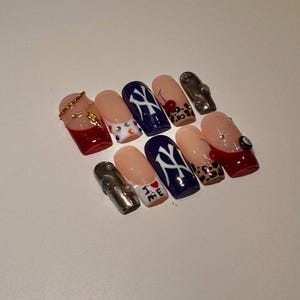 May include: A set of ten artificial nails with various designs. The nails feature a mix of colors, including red, navy blue, and nude. Some nails have gold accents, while others display patterns like a cherry, leopard print, and a stylized logo.