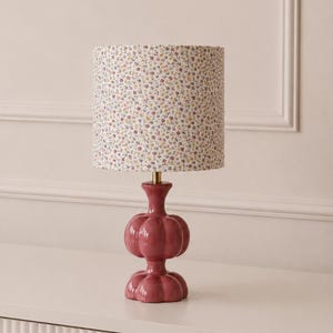 Può includere: Una lampada da tavolo con paralume cilindrico caratterizzato da un piccolo motivo floreale nei toni del rosa, giallo e blu. La base è in ceramica lucida color rosa scuro, con una forma impilata e arrotondata a zucca e un collo dorato.