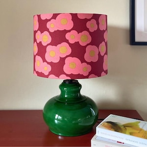 Può includere: Una lampada da tavolo in ceramica verde con base rotonda e paralume cilindrico. Il paralume presenta un motivo di fiori rosa e gialli su sfondo bordeaux. La lampada è su una superficie rossa.