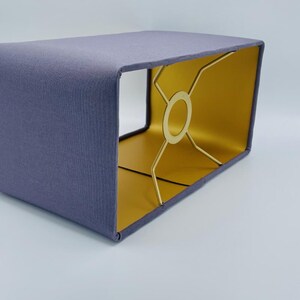 Könnte beinhalten: Ein rechteckiger Lampenschirm mit einer strukturierten, lavendelblauen Stoffaußenseite und einem glatten, goldfarbenen Innenfutter. Die Struktur wird von einem goldfarbenen Metallgestell mit einem zentralen Ring und vier Streben gehalten.