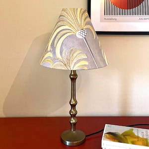 Puede incluir: Una lámpara de mesa de latón con una pantalla gris con un diseño de palmeras doradas y amarillas. La lámpara tiene una base delgada y decorativa y se encuentra sobre una superficie roja. Un libro es visible en la base de la lámpara.
