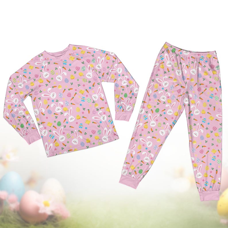 Op de afbeelding: Een roze tweedelige pyjamaset met een herhaald patroon van witte konijntjes, wortels, paaseieren en bloemen. De top met lange mouwen en de broek hebben roze manchetten.