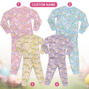 Op de afbeelding: Vier sets paaspyjama's. De pyjama's hebben tops en broeken met lange mouwen met patronen van konijnen, wortels en eieren. Kleuren zijn roze, blauw, geel en paars. De tekst "CUSTOM NAME" staat boven de pyjama's.