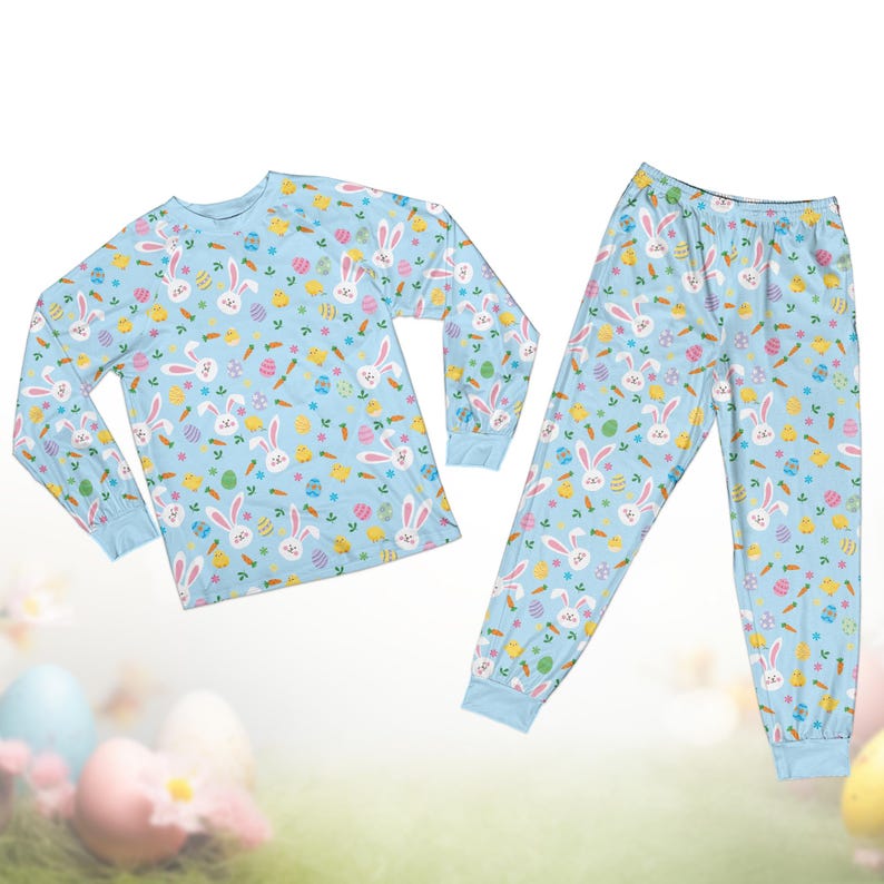 Op de afbeelding: Een lichtblauwe pyjamaset met een herhaald patroon van witte paashazen, kleurrijke eieren, wortels en kleine gele kuikens. De top met lange mouwen en de broek hebben lichtblauwe manchetten.