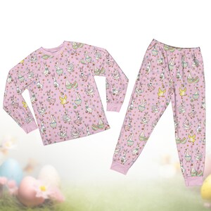 Op de afbeelding: Een lichtroze pyjamaset met een herhaald patroon van ganzen, konijnen en paaseieren. De top met lange mouwen en de broek hebben roze manchetten. De ganzen staan in verschillende poses, sommige dragen mutsen en houden manden vast.