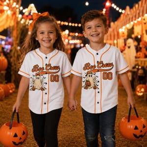 May include: Two white baseball jerseys with orange trim, featuring the text Boo Crew, the number 00, and a graphic of a ghost holding a pumpkin. The jerseys have a button-down front and short sleeves, worn with black pants and blue jeans.