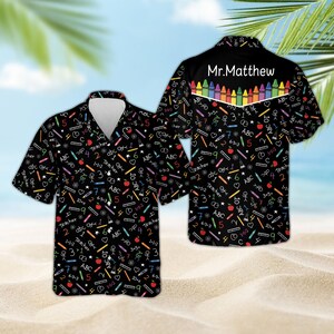May include: A black short-sleeved button-down shirt featuring a colorful pattern of school supplies, numbers, and math equations. The back yoke displays the text Mr. Matthew above a row of multi-colored crayons. The shirt has a notched collar.