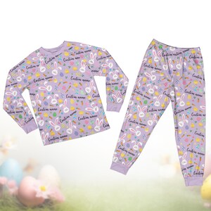 Op de afbeelding: Een lila pyjamaset met een top met lange mouwen en een broek. De set is bedekt met een patroon van witte paashazen, wortels, kuikens en versierde eieren. De tekst "Custom name" wordt herhaald.