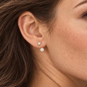 May include: A double-sided earring featuring two round, clear, faceted stones. The smaller stone sits directly on the earlobe, while the larger stone hangs slightly below the lobe, creating a layered, suspended appearance with a metallic setting.