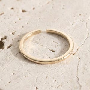 May include: A simple, thin, gold-colored metal ring with an open band design. The ring features a smooth, polished surface and a circular shape with a small gap between the two ends of the band, creating an adjustable style.