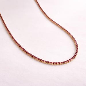 Może przedstawiać: Delikatny złoty naszyjnik z ciągłą linią małych, kwadratowych, ciemnoczerwonych kamieni szlachetnych. Naszyjnik jest prezentowany na miękkim, białym tle, podkreślając żywy kolor i szczegółowy design biżuterii.