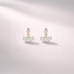May include: A pair of gold-colored earrings with a unique design. Each earring features a small, round gem at the top and a cluster of marquise-cut gems at the bottom. The earrings are set against a soft, light pink background.