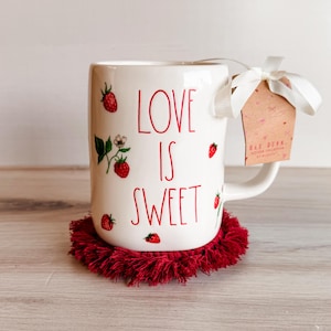 Peut inclure: Mug en céramique blanche avec l'inscription rouge « LOVE IS SWEET », décorée d'illustrations de fraises. Le mug est posé sur un dessous de verre rouge à franges et porte une étiquette attachée avec un ruban blanc.