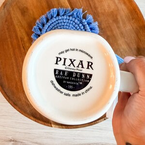K&ouml;nnte beinhalten: Die Unterseite einer wei&szlig;en Keramiktasse mit schwarzem Text, der PIXAR, RAE DUNN, ARTISAN COLLECTION BY MAGENTA, 195, may get hot in microwave und dishwasher safe, made in china liest. Ein hellblauer, runder Makramee-Untersetzer ist dahinter zu sehen.