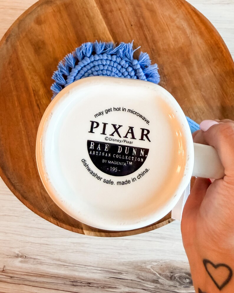 K&ouml;nnte beinhalten: Die Unterseite einer wei&szlig;en Keramiktasse mit schwarzem Text, der PIXAR, RAE DUNN, ARTISAN COLLECTION BY MAGENTA, 195, may get hot in microwave und dishwasher safe, made in china liest. Ein hellblauer, runder Makramee-Untersetzer ist dahinter zu sehen.