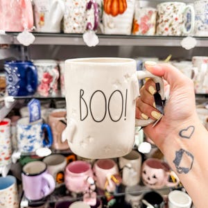 May include: A white ceramic mug with the word "BOO!" in black lettering. The mug has a ghost-shaped handle and star details. The background shows a variety of other mugs on shelves.