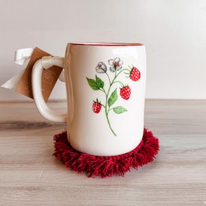Peut inclure: Mug en céramique blanche avec un bord rouge et une anse, orné d'une illustration à l'aquarelle de framboises et de fleurs. Le mug est posé sur un dessous de verre rond rouge à franges. Une étiquette est attachée à l'anse avec un ruban blanc.