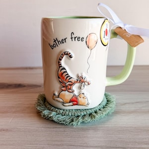 May include: A cream-colored ceramic mug featuring a raised illustration of Tigger standing on Winnie the Pooh, with the text "bother free" and a balloon. The mug has a light green interior and handle, with a white ribbon tied to the handle.