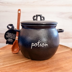 May include: A black, round, textured ceramic cauldron with a matching lid and two side handles. The word potion is written in white script on the front. A brown wooden spoon is tucked behind the cauldron, and a black tag is attached to the handle.