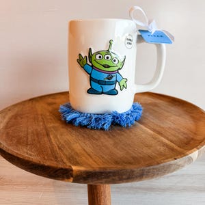K&ouml;nnte beinhalten: Ein wei&szlig;er Keramikbecher mit einem gr&uuml;nen, drei&auml;ugigen Alien in einem blauen Raumanzug. Am Henkel befindet sich ein wei&szlig;es Band mit einem blauen Pixar-Etikett. Der Becher steht auf einem runden, blauen Untersetzer mit Fransen und einem Aufkleber.