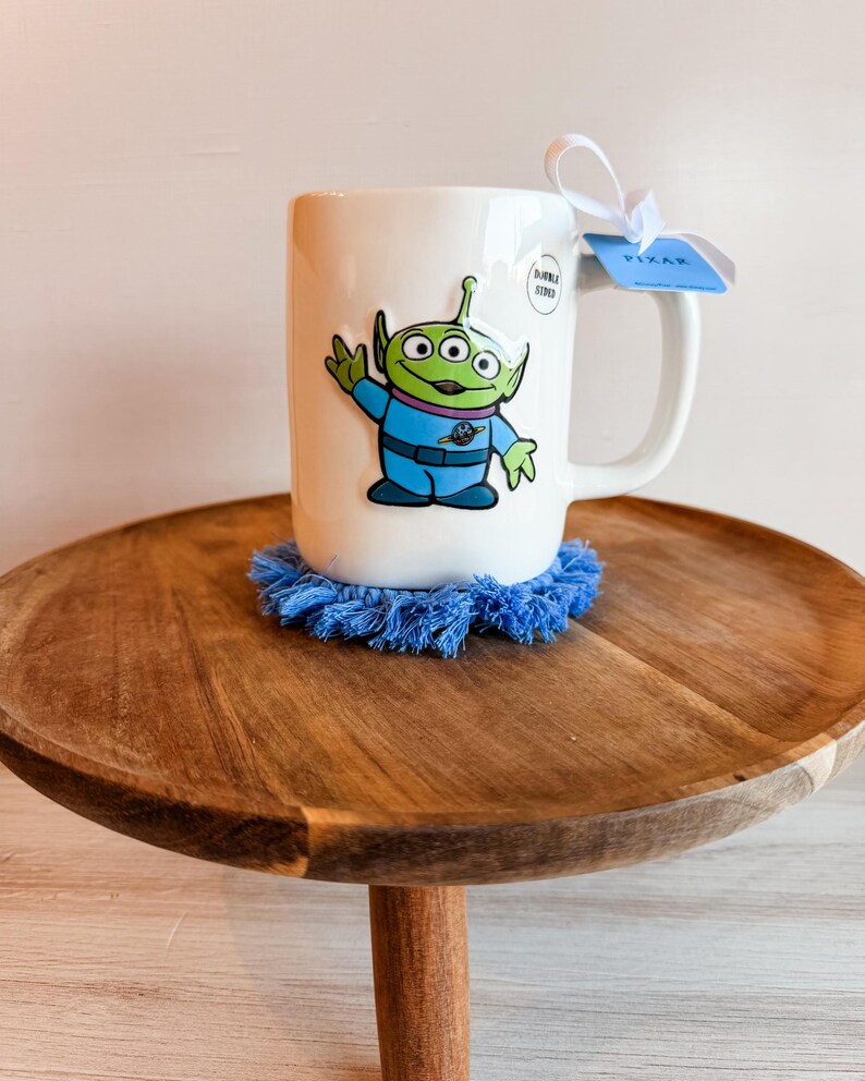 K&ouml;nnte beinhalten: Ein wei&szlig;er Keramikbecher mit einem gr&uuml;nen, drei&auml;ugigen Alien in einem blauen Raumanzug. Am Henkel befindet sich ein wei&szlig;es Band mit einem blauen Pixar-Etikett. Der Becher steht auf einem runden, blauen Untersetzer mit Fransen und einem Aufkleber.