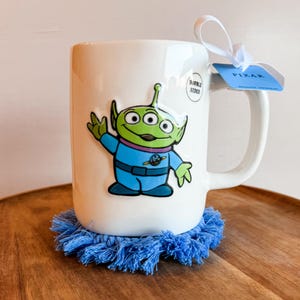 K&ouml;nnte beinhalten: Ein wei&szlig;er Keramikbecher mit einem gr&uuml;nen, drei&auml;ugigen Alien in einem blauen Raumanzug. Am Henkel befindet sich ein wei&szlig;es Band mit einem blauen Etikett mit der Aufschrift PIXAR. Ein kleiner runder Aufkleber zeigt DOUBLE SIDED. Der Becher steht auf einem blauen Fransenuntersetzer.