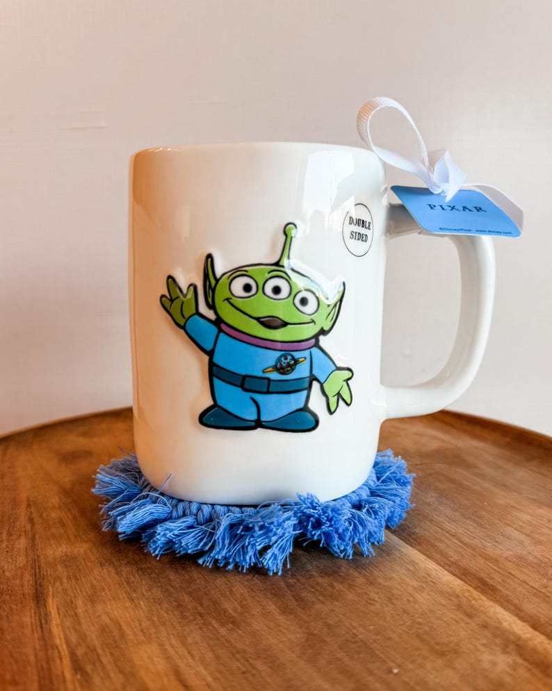 K&ouml;nnte beinhalten: Ein wei&szlig;er Keramikbecher mit einem gr&uuml;nen, drei&auml;ugigen Alien in einem blauen Raumanzug. Am Henkel befindet sich ein wei&szlig;es Band mit einem blauen Etikett mit der Aufschrift PIXAR. Ein kleiner runder Aufkleber zeigt DOUBLE SIDED. Der Becher steht auf einem blauen Fransenuntersetzer.