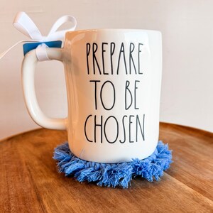 K&ouml;nnte beinhalten: Ein wei&szlig;er Keramikbecher mit dem schwarzen Text PREPARE TO BE CHOSEN auf der Vorderseite. Ein wei&szlig;es Band ist am Henkel befestigt. Der Becher steht auf einem runden, blauen Makramee-Untersetzer mit Fransen. Das Design wirkt schlicht und klar.