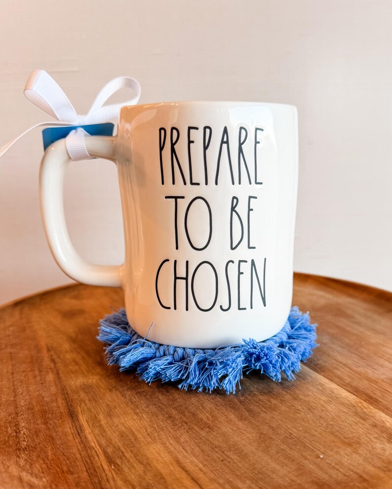 K&ouml;nnte beinhalten: Ein wei&szlig;er Keramikbecher mit dem schwarzen Text PREPARE TO BE CHOSEN auf der Vorderseite. Ein wei&szlig;es Band ist am Henkel befestigt. Der Becher steht auf einem runden, blauen Makramee-Untersetzer mit Fransen. Das Design wirkt schlicht und klar.