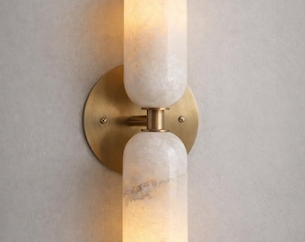 May include: A contemporary wall light with two cylindrical alabaster shades in off-white. The shades are mounted on a circular, brass-toned backplate. This light fixture is designed for indoor use and adds a touch of elegance to any room.