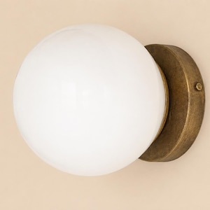 May include: A round, white glass globe light fixture with a brass-colored metal base. The fixture is mounted on a beige wall. The globe is smooth and opaque, and the base has a circular design.