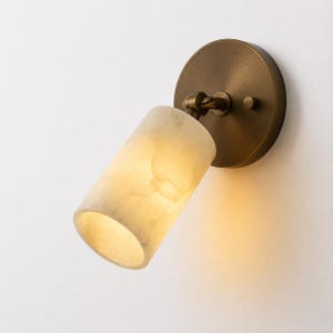 May include: A wall-mounted light fixture with a cylindrical, off-white shade and a circular, brass-colored base. The shade is angled, and the fixture is designed to direct light. The fixture is mounted on a white wall.