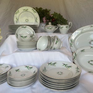 May include: A complete vintage porcelain dinnerware set. The white dishes feature a green leaf design and gold trim. The set includes plates, bowls, cups, saucers, serving platters, a sugar bowl, and a creamer.