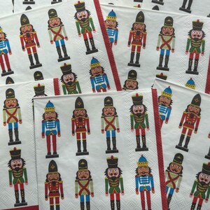 May include: White paper napkins with a repeating pattern of colorful nutcracker soldiers. The nutcrackers are depicted in various uniforms, including red, blue, and green, with black boots and hats. Red stripes border some of the napkins.