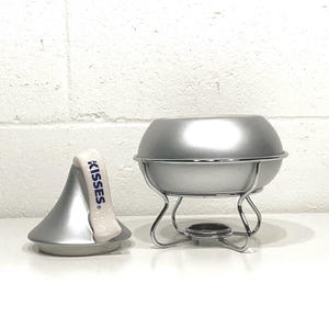 May include: A silver-colored fondue pot with a matching lid and stand. The pot sits on a chrome-colored stand with curved legs. A silver-colored Hershey's Kisses-shaped lid is next to the pot, with the word "KISSES" printed on it.