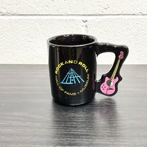 Op de afbeelding: Zwarte keramische mok met het logo van het Rock and Roll Hall of Fame Museum in blauw en geel. De handgreep heeft de vorm van een roze gitaar. De mok staat op een donkere ondergrond.
