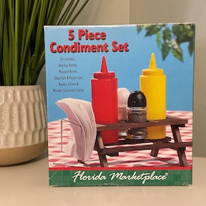 Pode incluir: Um conjunto de condimentos de 5 peças com garrafas de ketchup e mostarda, um conjunto de sal e pimenta e um porta-guardanapos. O conjunto é apresentado numa pequena mesa de piquenique de madeira. A caixa diz "Florida Marketplace."