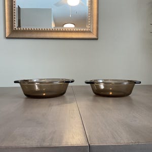 May include: Two brown glass bowls with black handles are displayed on a light brown wooden table. The bowls are oval-shaped and appear to be oven-safe. A gold-framed mirror hangs in the background.