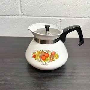 Puede incluir: Tetera de esmalte blanco con asa y tapa negras. La tetera presenta una banda decorativa y un diseño de frutas y flores. El texto "Le Thé" está impreso en la parte delantera.