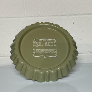 May include: An olive green flan pan with a scalloped edge. The pan has a white printed recipe for a basic flan. The pan is made of metal and is sitting on a white surface.