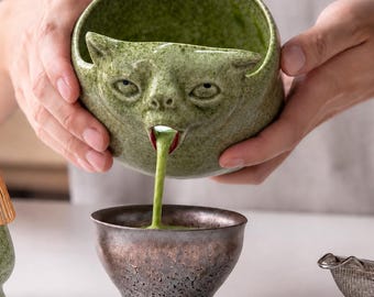 May include: A green speckled ceramic bowl shaped like a cat head with an open mouth pouring green matcha tea into a smaller metallic-finished bowl. The cat-shaped bowl features sculpted ears, eyes, and a nose, with a dark red accent inside the mouth.