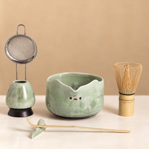 May include: A matcha tea set featuring a light green ceramic bowl with a small bird design, a matching whisk holder with a metal mesh strainer, a bamboo whisk, and a thin bamboo tea scoop resting on a small ceramic bird-shaped rest.