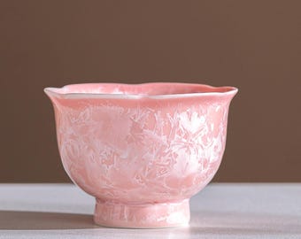May include: A pink ceramic bowl with a unique crystalline glaze. The bowl has a rounded body, a flared rim, and a small, pedestal base. The glaze has a textured, almost frosted appearance, with white and pink hues. The bowl is likely used for serving or display.