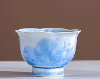 May include: A white and blue ceramic teacup with a unique crystalline pattern. The cup has a flared rim and a pedestal base. The blue design covers the lower portion of the cup, creating a striking contrast against the white background. The teacup is a decorative item.