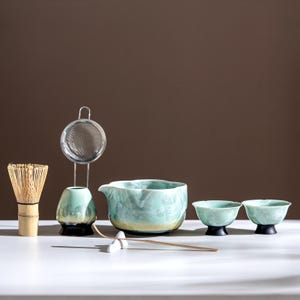 May include: A matcha tea set comprising a bamboo whisk, a stainless steel sieve, a ceramic bowl, two small cups, and a ceramic water container. The set has a light blue and cream colour scheme, with black accents on the cups and water container.