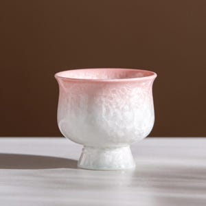 Pode incluir: Uma tigela de cerâmica com pé e gradiente de cores. A borda é rosa suave, desbotando para branco com um padrão cristalino texturizado. A base é verde pálido e a tigela assenta sobre um pé curto e alargado.