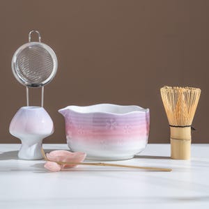 May include: A matcha tea set featuring a stainless steel strainer, a ceramic bowl with a pink and purple gradient, a bamboo whisk, and a ceramic spoon. The bowl has a floral design. The set is arranged on a white surface.
